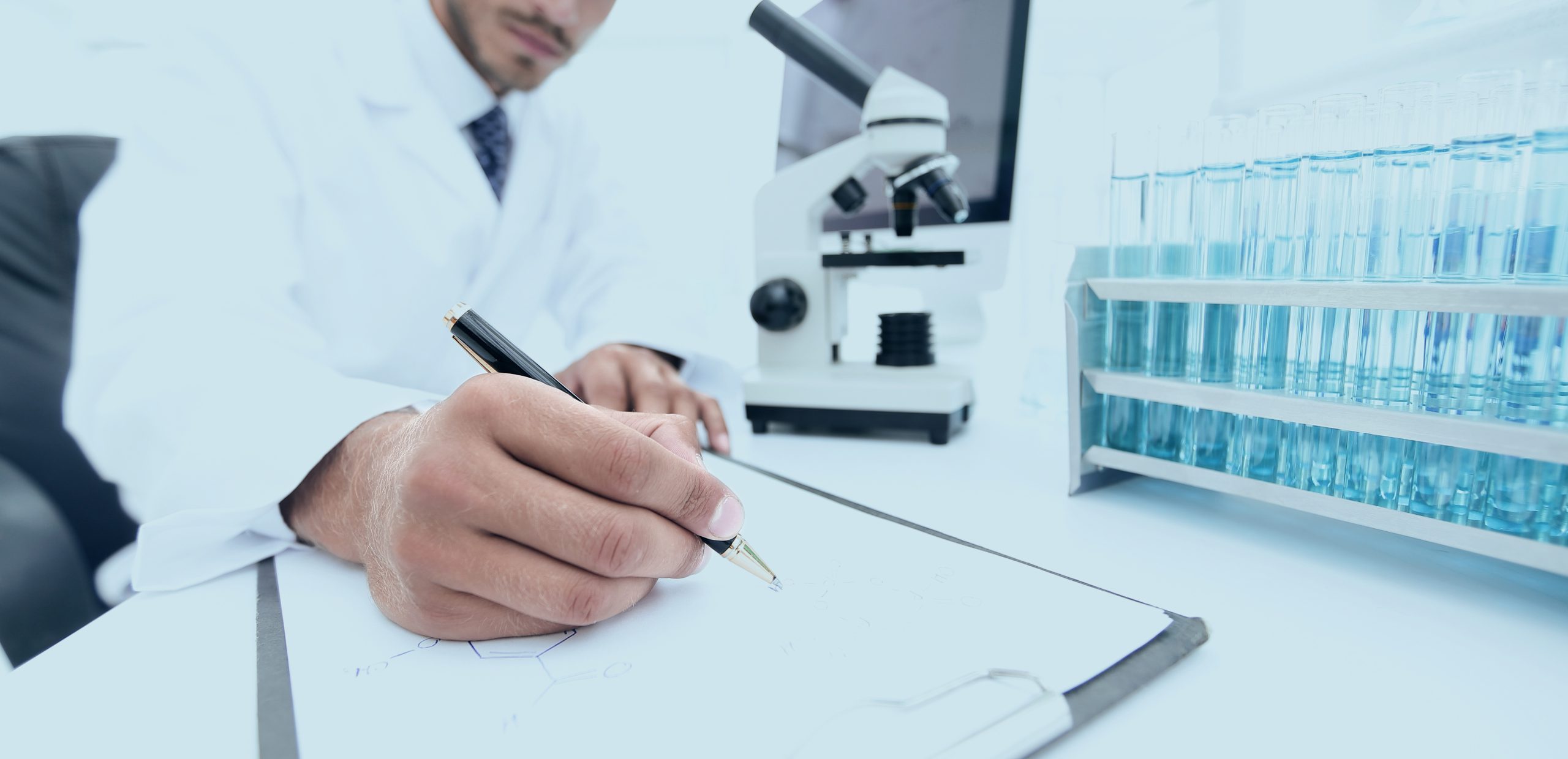 Fotografia de cientista branco com cavanhaque escrevendo em laboratório. Ele está sentado a uma mesa, com um microscópio e tubos de ensaio na sua frente