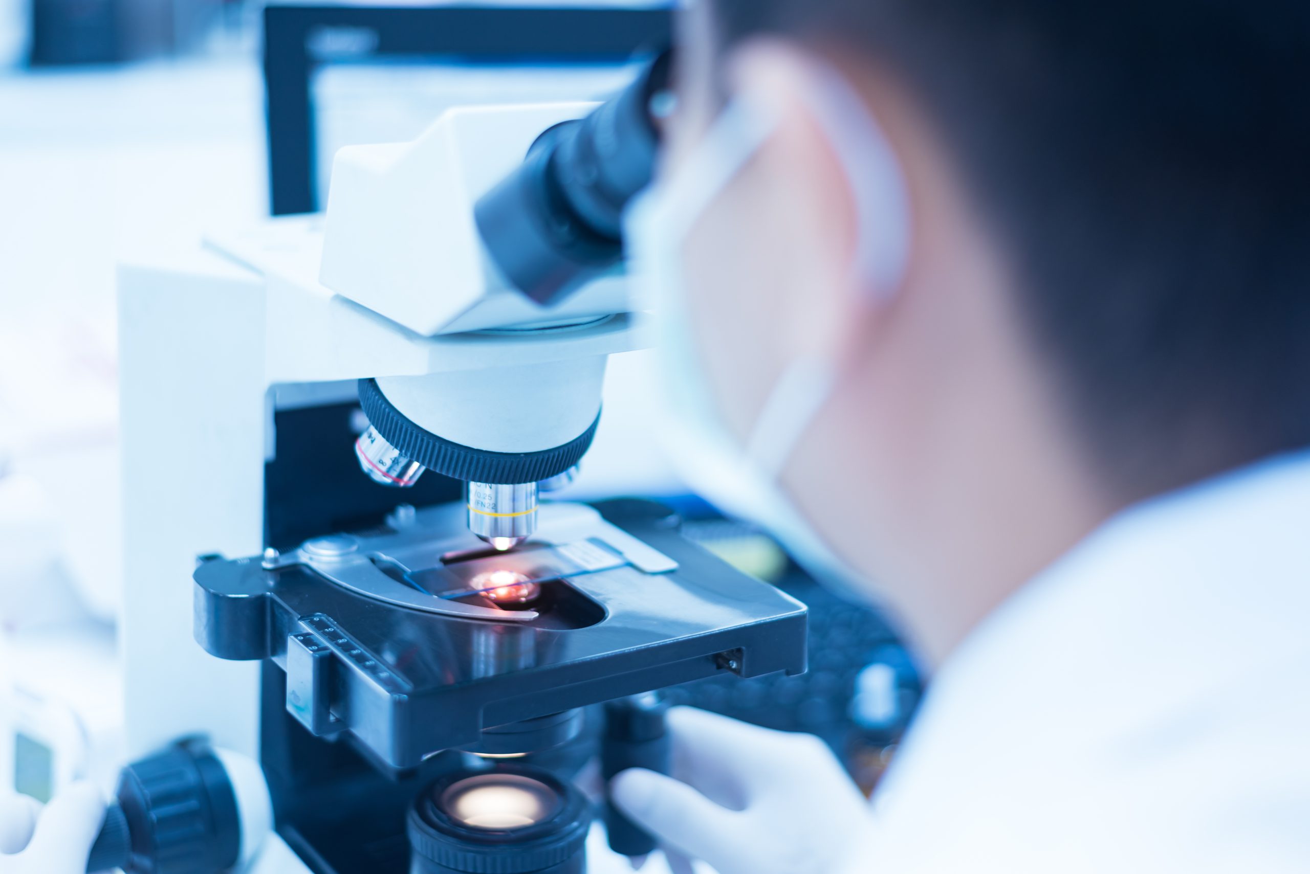 Fotografia com foco seletivo de microscópio, com parte do rosto de cientista a frente.