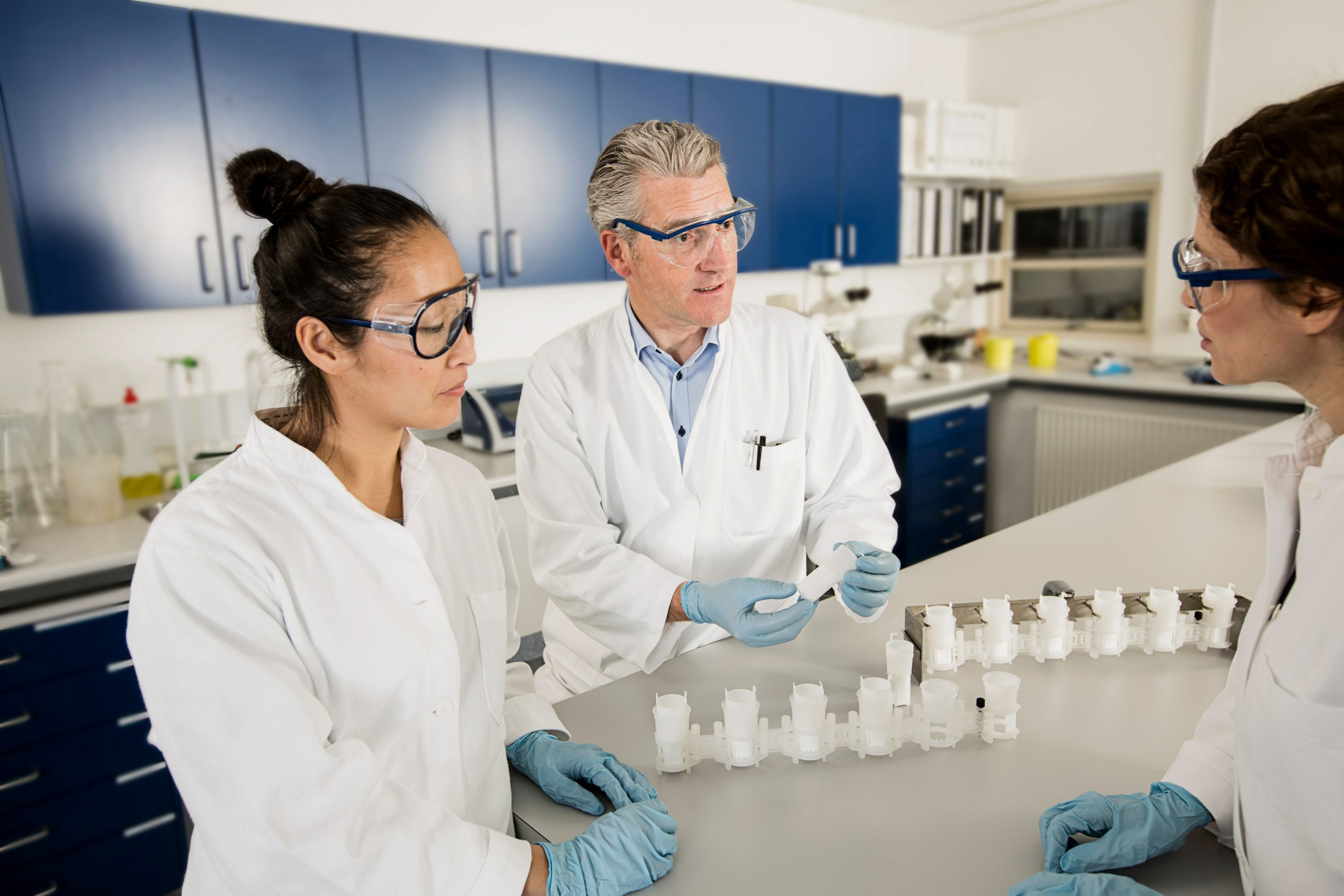 Fotografia de grupo de tr^ss cientistas em laboratório conversando. São duas mulheres brancas e um homem também branco, todos estão usando jaleco branco e óculos de proteção.