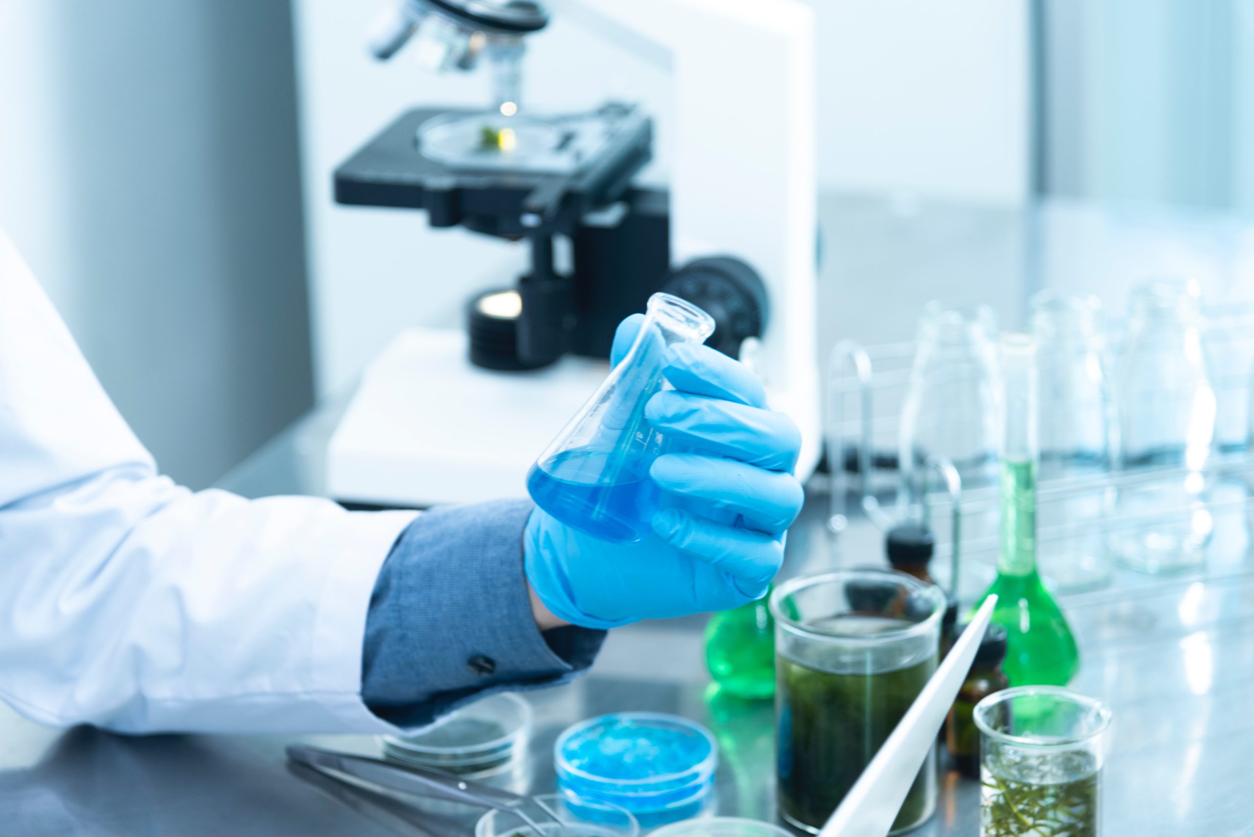 Fotografia de balcão de laboratório com mão usando luva segurando erlenmeyer pequeno com um líquido azul