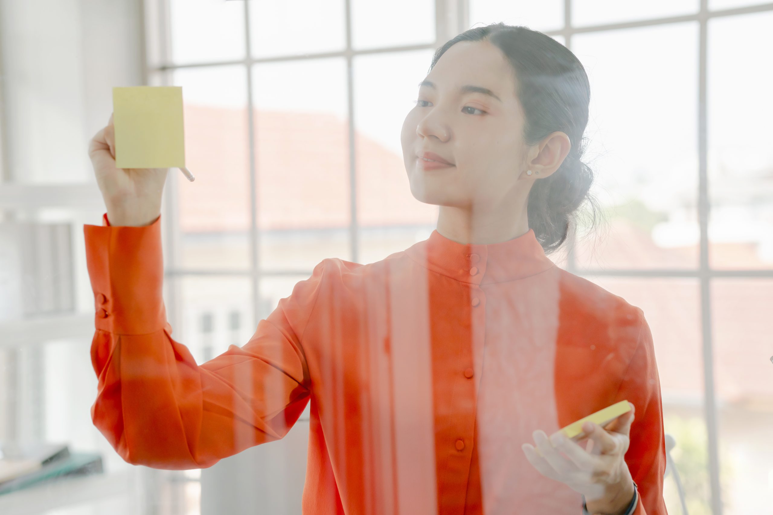 Fotografia de mulher asiática escrevendo em post-it colado em vidro