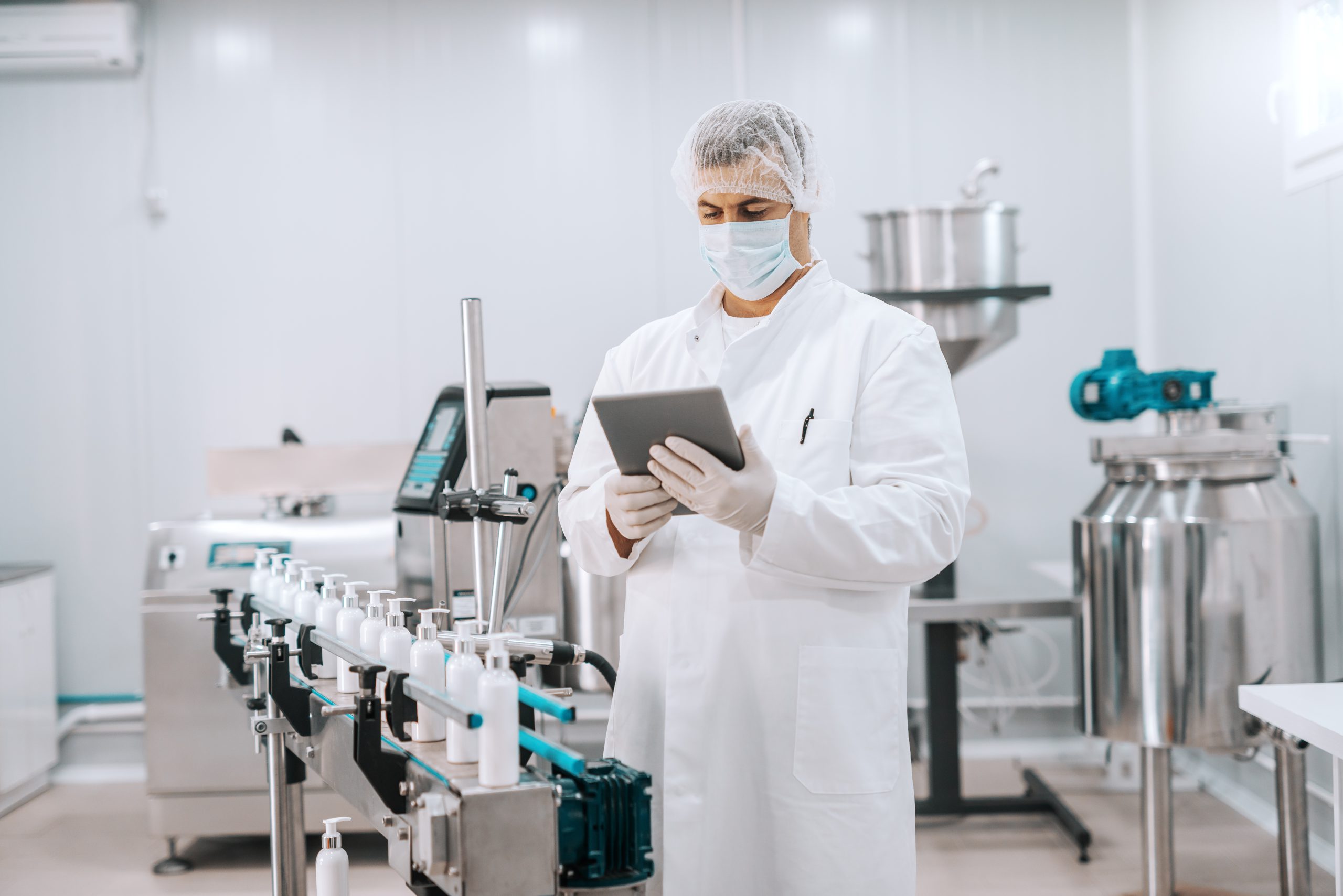 Fotografia de homem branco analisando cadeia de produção em laboratório. Ele usa jaleco, luvas, touca e máscara e olha para um tablet com expressão concentrada. O homem está no interior de uma fábrica química.