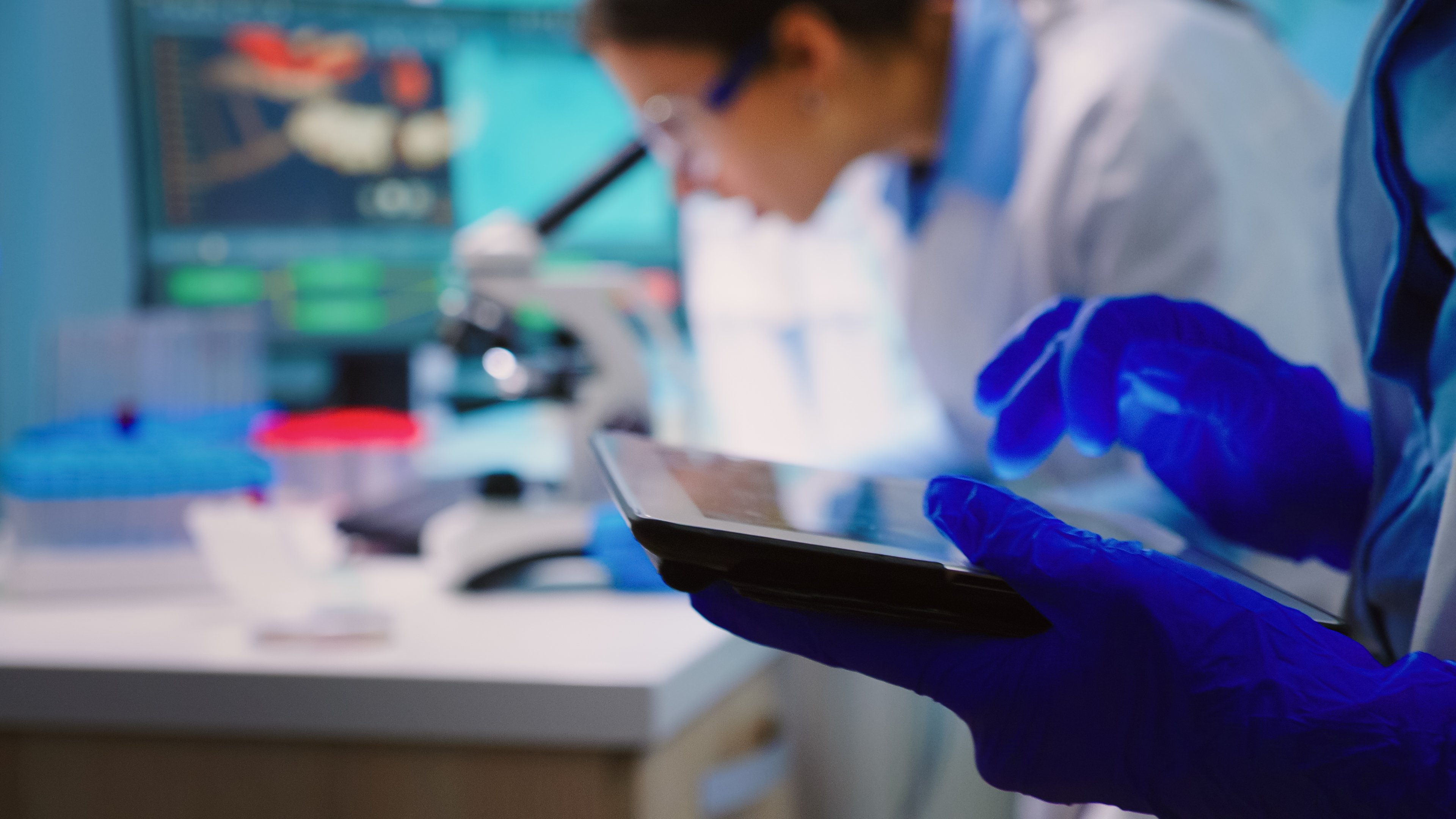 close-up-scientist-writing-tablet-while-team-biologists-doing-biological-researches-microscope-backgorund-late-night