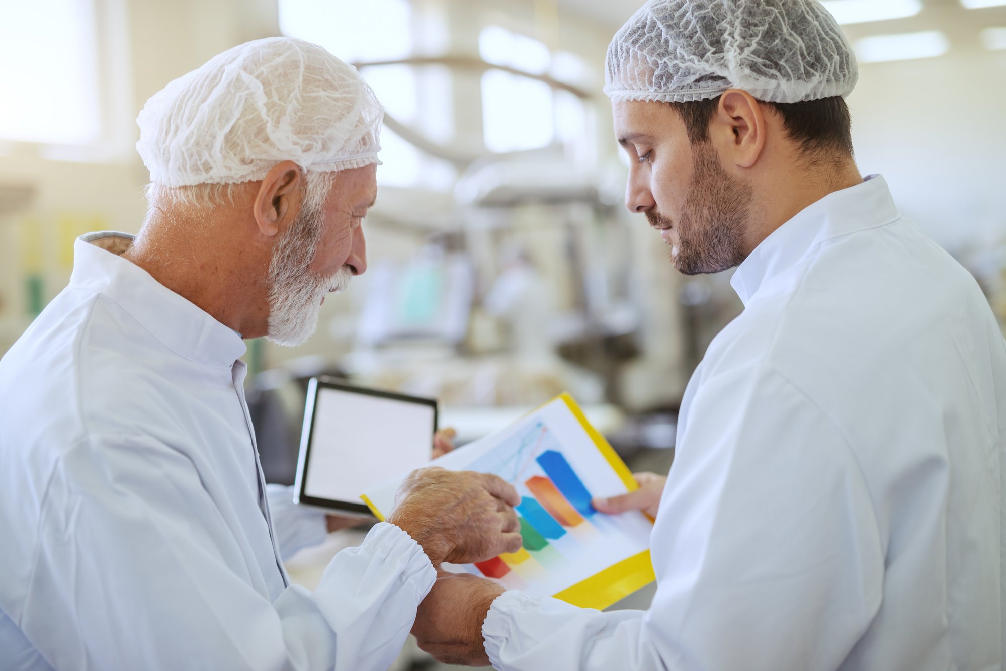 two-hardworking-supervisors-checking-statistics-while-standing-food-plant-older-one-holding-tablet-both-are-dressed-white-sterile-uniforms-1