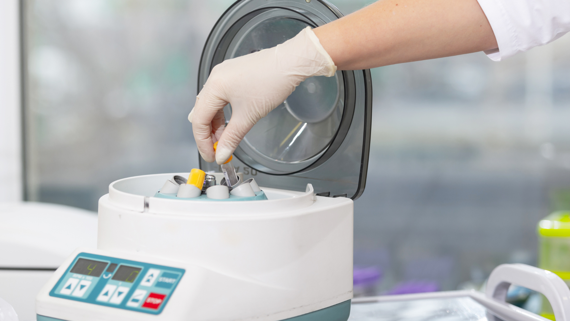 Fotografia de centrífuga em laboratório, com uma mão branca usando luva inserindo uma amostra na máquina