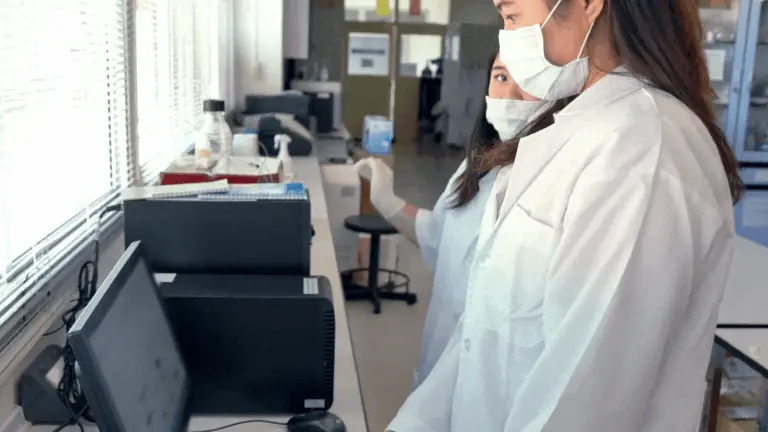 Duas analistas mascaradas observando equipamento de laboratório, validação de métodos analíticos.